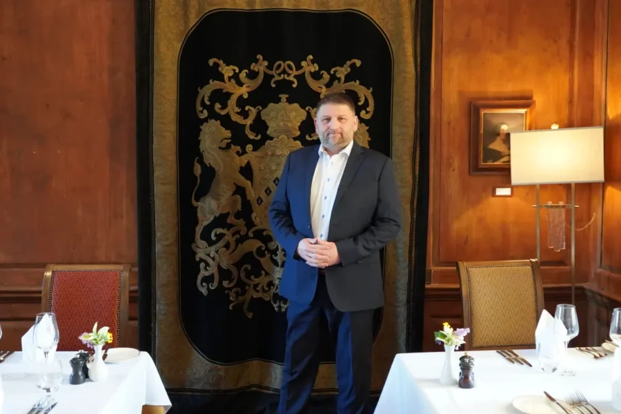 Richard Stone in a panelled dining room standing in front of a heraldic tapestry, navy suit.
